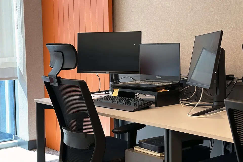 Loop Games Ofis çalışma istasyonu detayı, çift monitör düzeni, ergonomik sandalye ve turuncu dikey lata duvar paneli