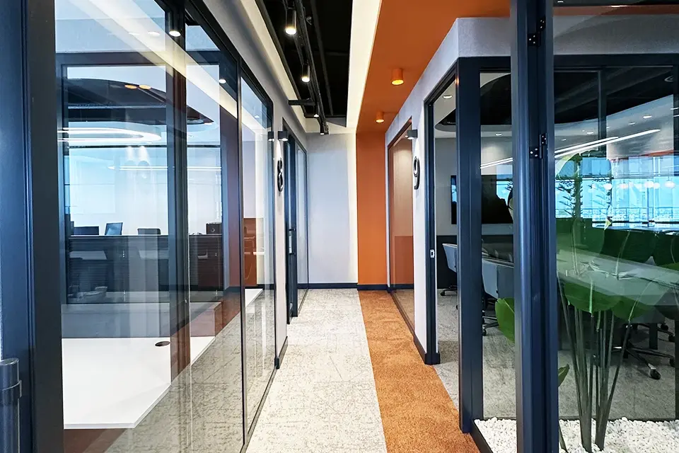 Loop Games Ofis cam toplantı odası koridoru, turuncu halı aksı, bitki terraryumları, siyah çerçeve cam bölmeler