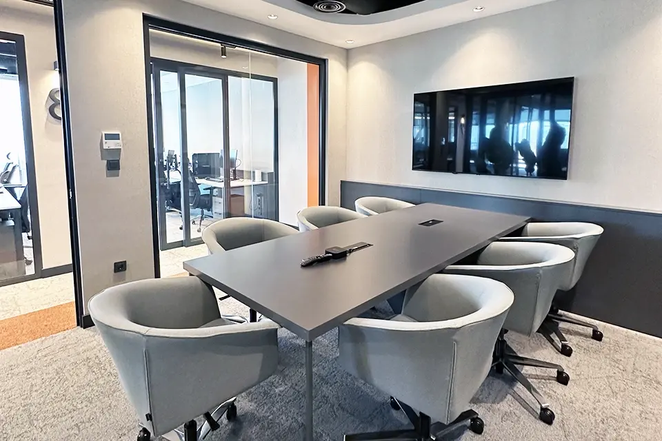 Loop Games Ofis toplantı odası, koyu gri toplantı masası, gri kumaş sandalyeler, duvar montaj TV ve cam sürgülü kapı