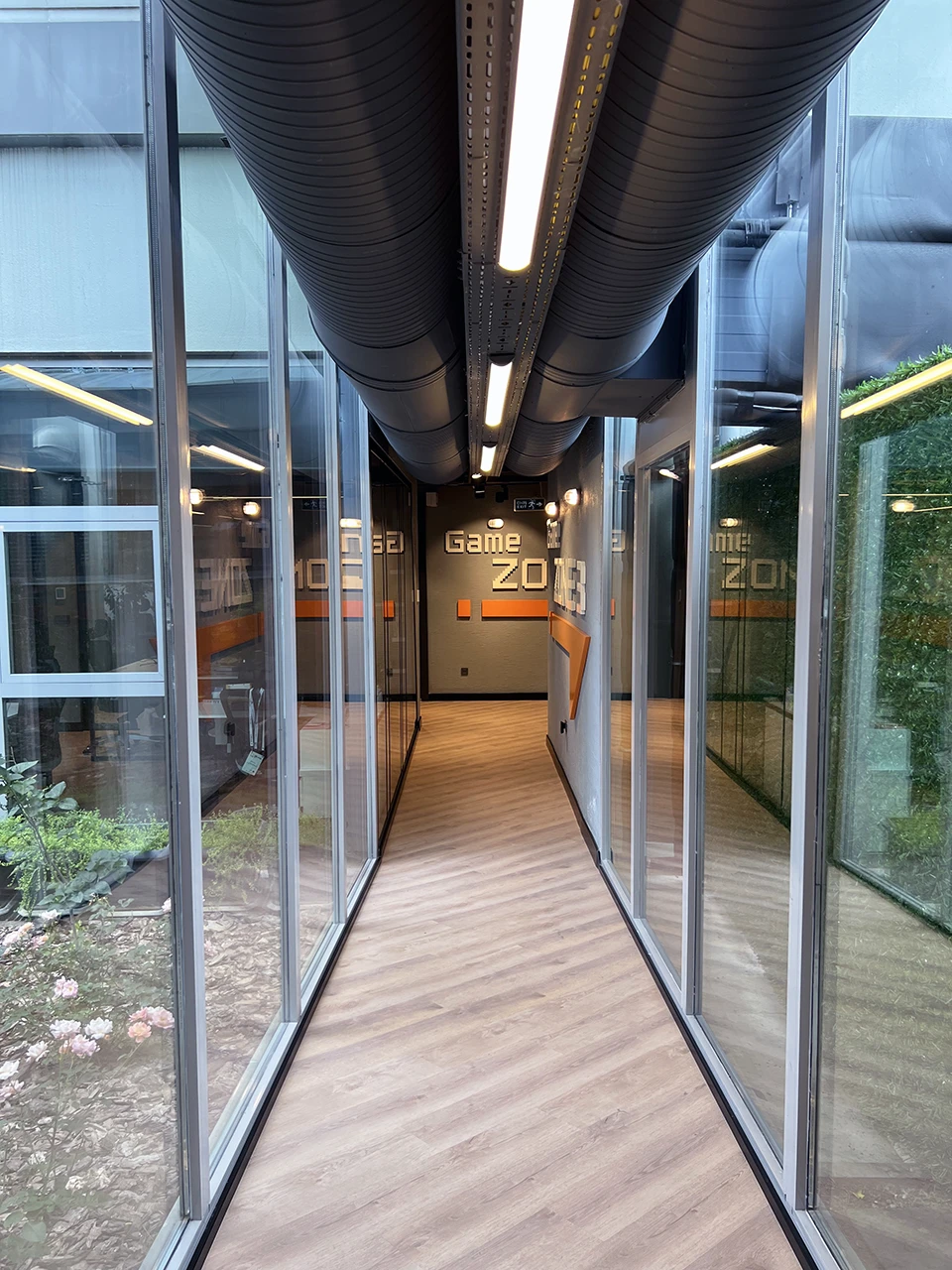 Panteon Ofis cam koridor geçiş alanı, iki taraflı cam cephe, iç bahçe manzarası, Game Zone 3 yazısı ve endüstriyel havalandırma